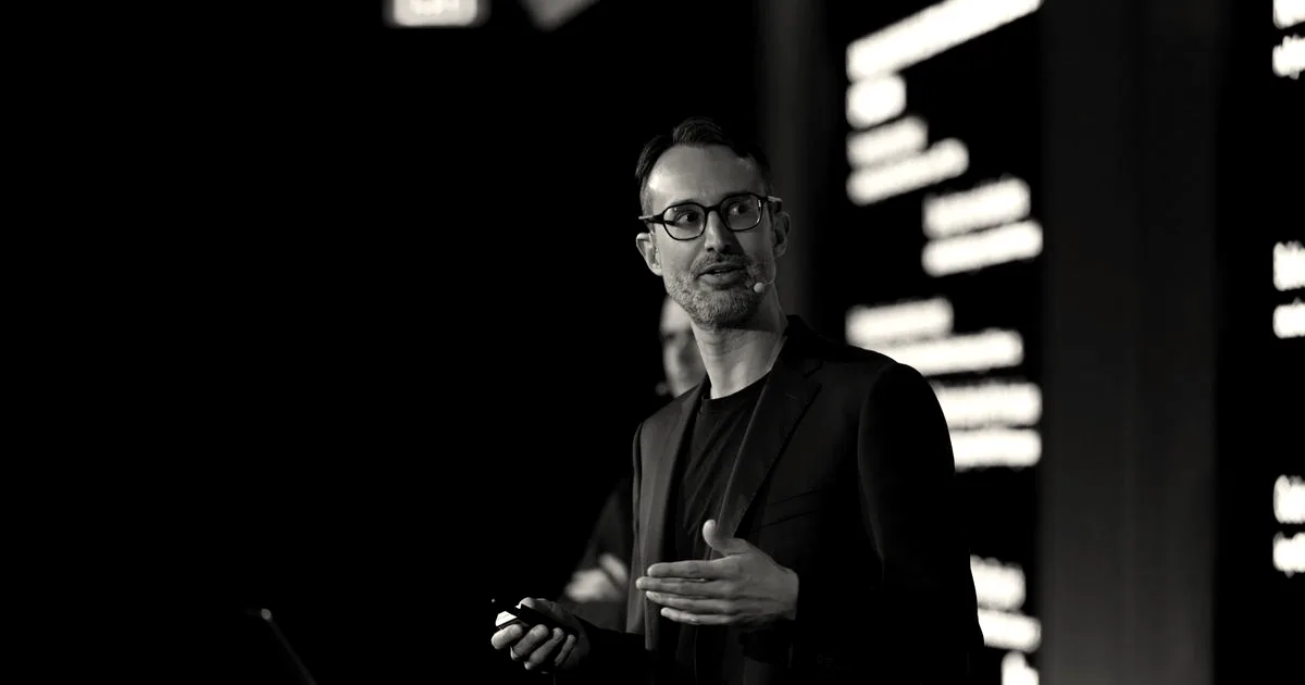 Kog CEO Gaël Dellalleau speaking on stage in front of presentation slides wearing glasses and a suit jacket.