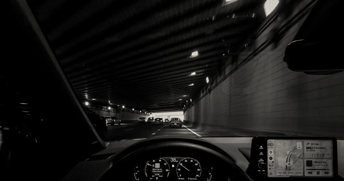 View from car dashboard driving through an illuminated city tunnel at night.