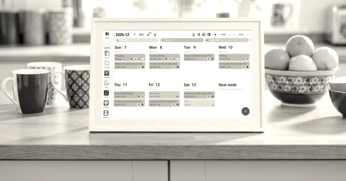 A white digital calendar display sitting on a wooden kitchen counter alongside mugs and fruit.