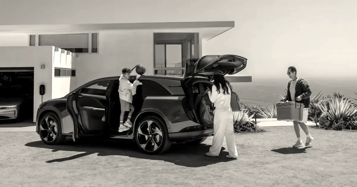 A family unloads the trunk and doors of a dark Lucid electric SUV parked outside a modern home.