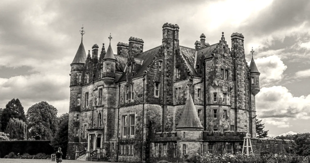 A grand historic stone castle with spires and manicured gardens under a cloudy blue sky.