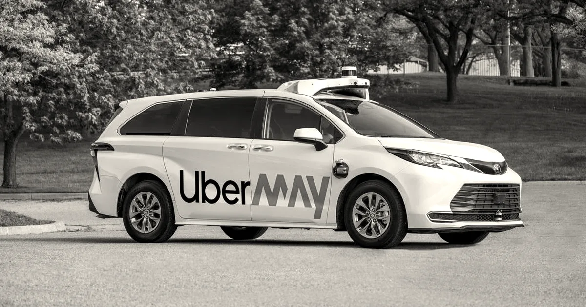A white autonomous May Mobility and Uber branded minivan parked outdoors on asphalt near green trees.