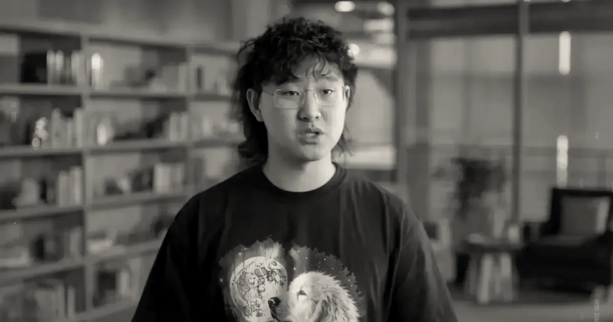 A young man with glasses and curly hair wearing a dog graphic t-shirt in front of a bookshelf.