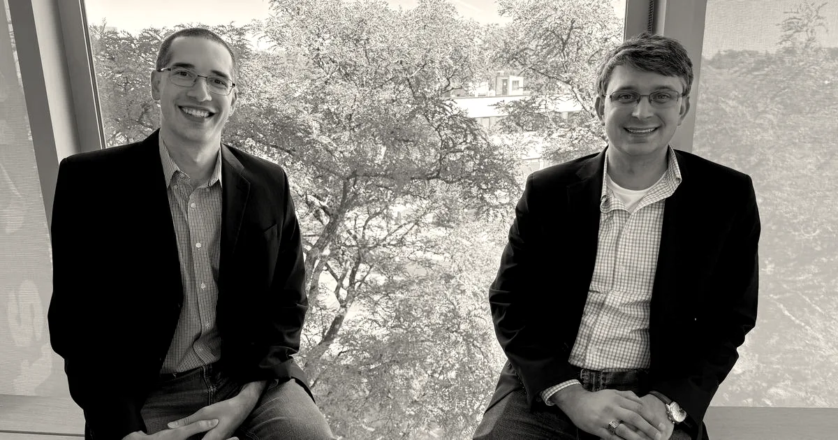 Two smiling cofounders in blazers and jeans sitting in front of a bright window overlooking green trees.