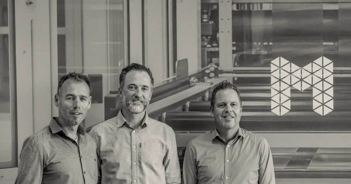 Three company leaders standing side-by-side in front of a blue background with a geometric M logo.
