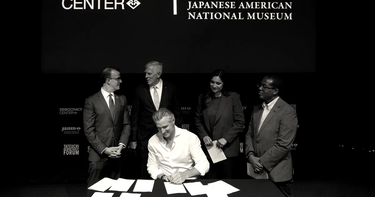 California Governor Gavin Newsom sits at a desk signing legislation surrounded by officials on a stage.