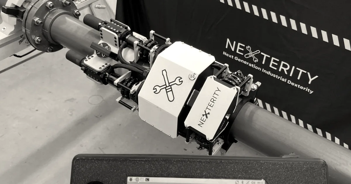 A handheld controller displaying the Nexterity logo held in front of a robotic pipefitting device on a pipe.