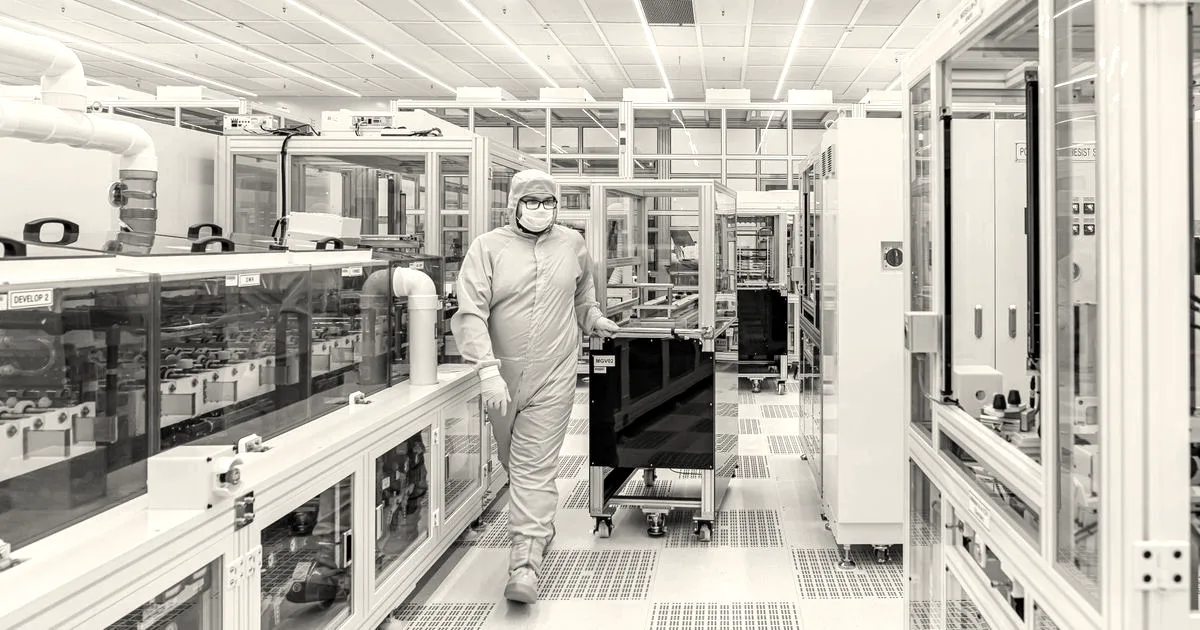 A technician wearing a white cleanroom suit walks through a bright high-tech semiconductor fabrication laboratory.