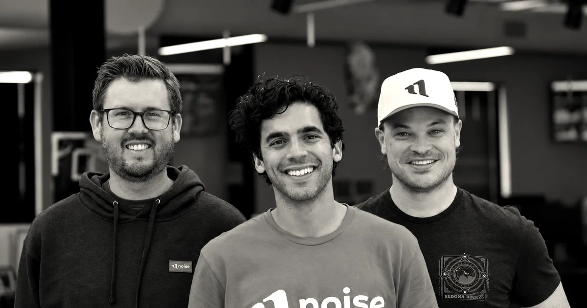 Three team members smiling together indoors, with the central man wearing a red t-shirt reading Noise.