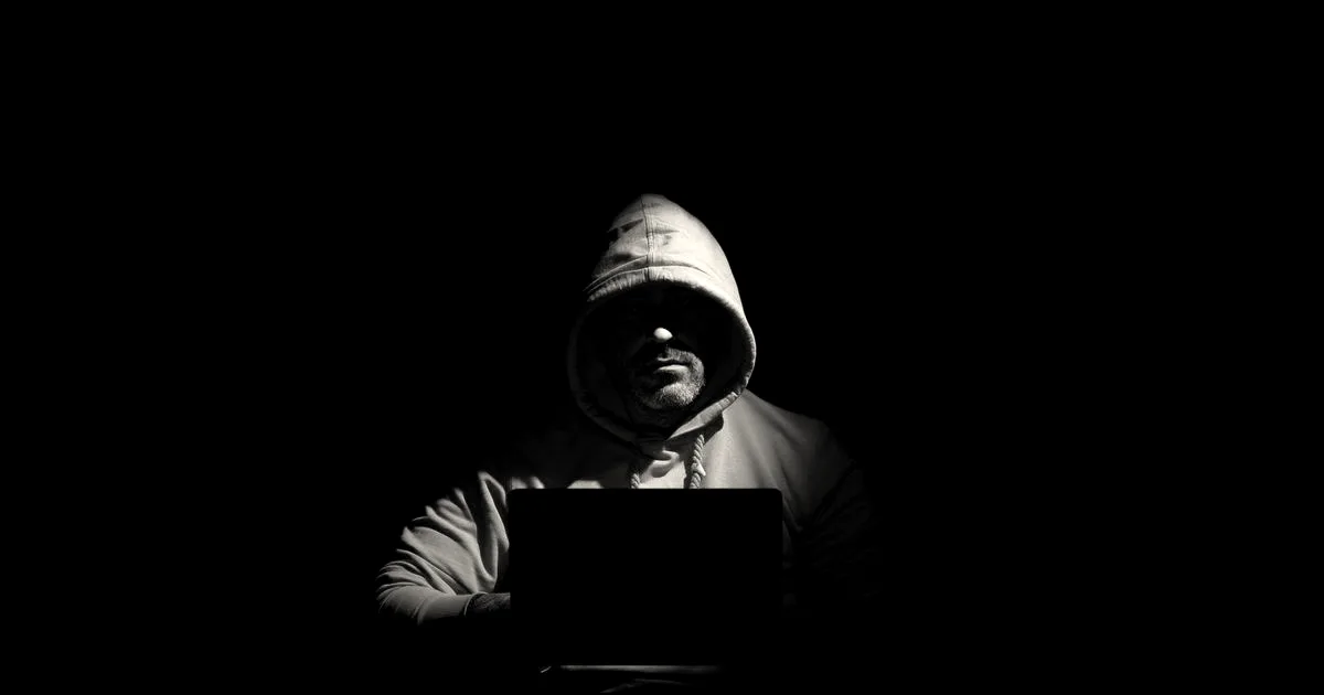 A hooded person sits in darkness illuminated only by the screen of an open laptop.