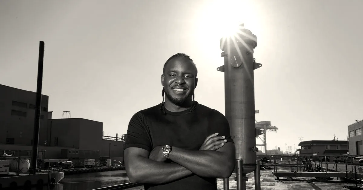 Man smiling with arms crossed outdoors next to industrial nuclear reactor hardware under bright sunlight.
