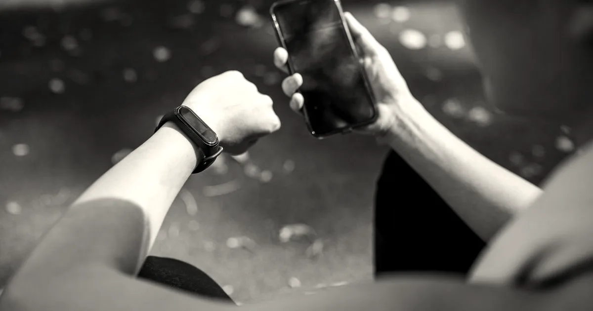 A person checking a fitness tracker wristband while holding a smartphone in their other hand