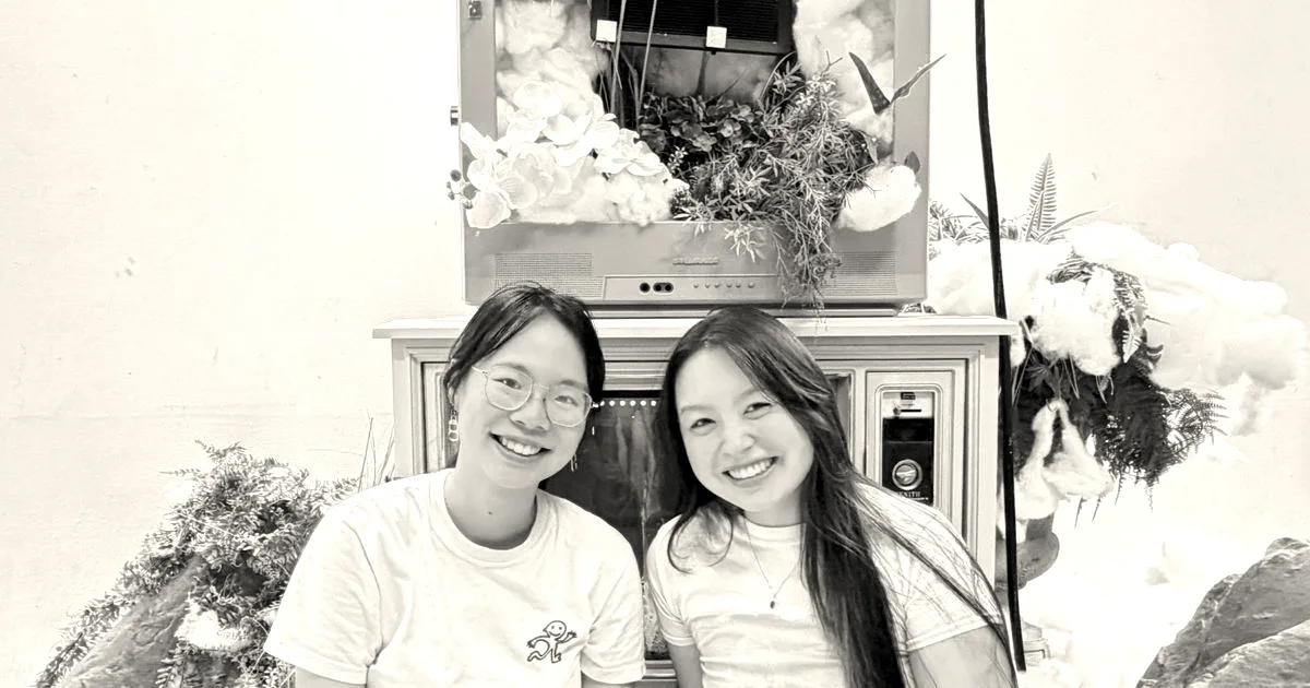 Two young women smiling in front of a decorative pastel television set adorned with flowers and cotton clouds.