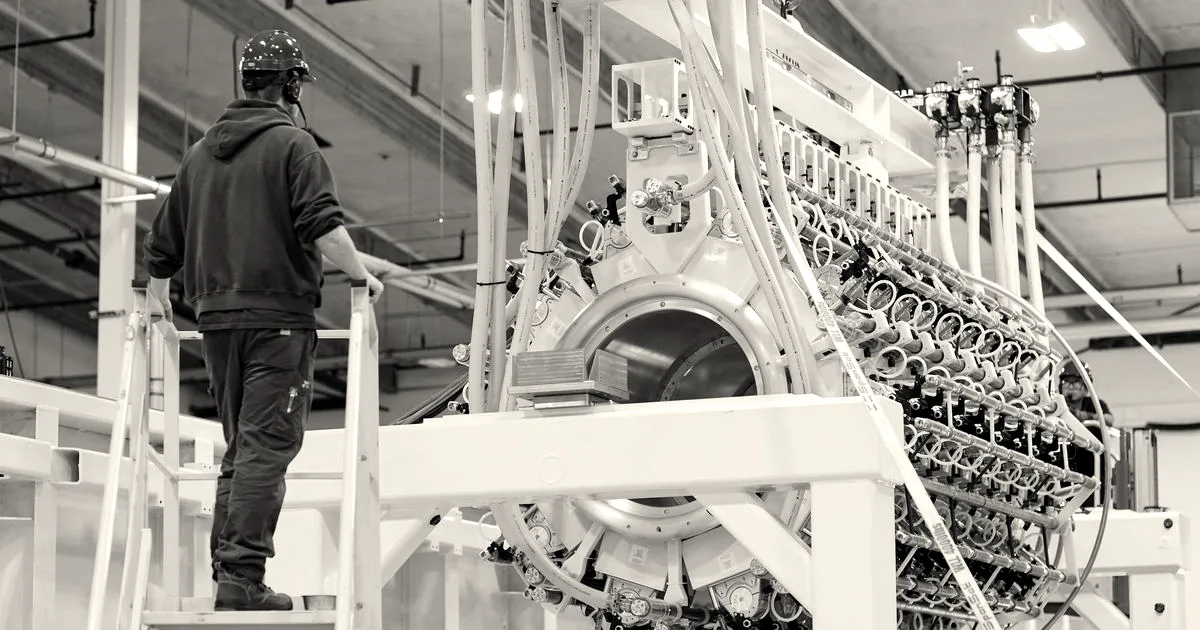 An engineer stands on a yellow ladder inspecting a large cylindrical fusion pulser prototype in a facility.
