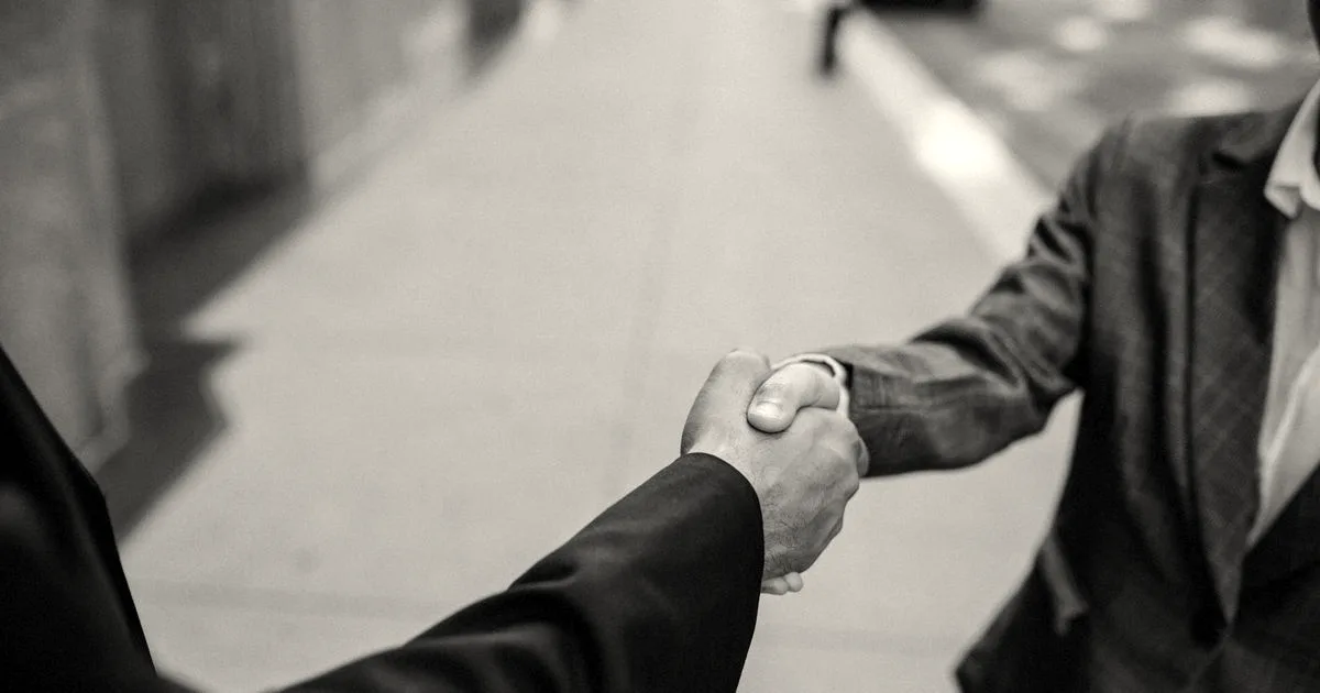 Two business professionals wearing suits shake hands outdoors, symbolizing a completed corporate deal or partnership.