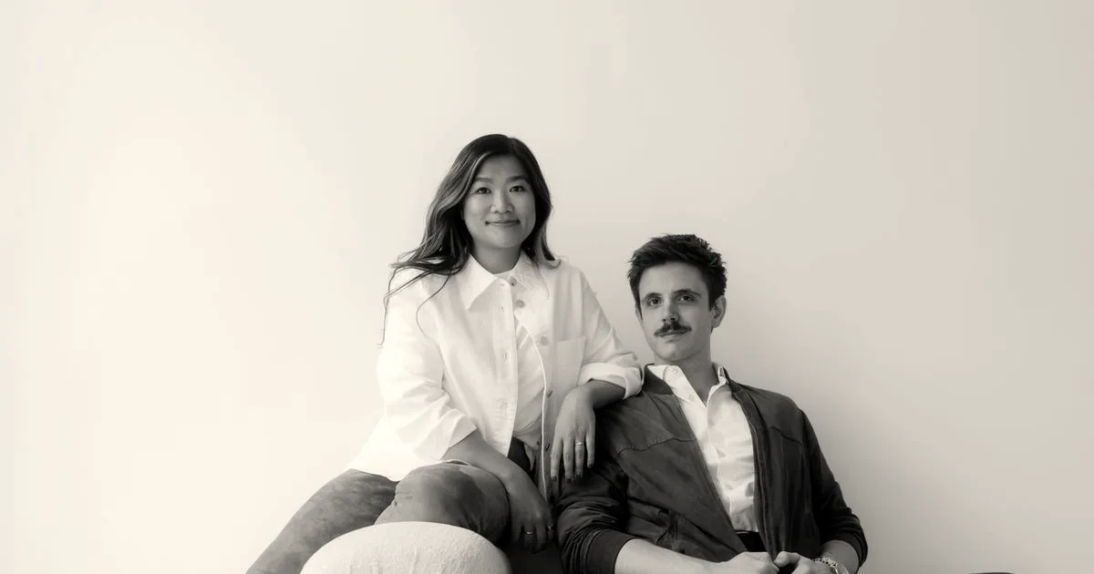 A man and woman sitting together on a light-colored couch against a white wall.