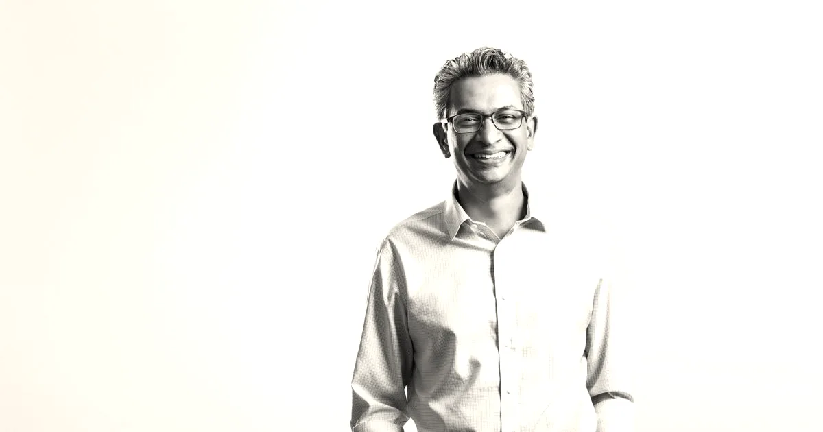 Smiling man in a light blue button-down shirt and glasses against a light background.