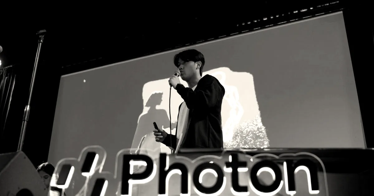 A presenter speaking on stage behind a glowing Photon sign with a presentation screen in the background.