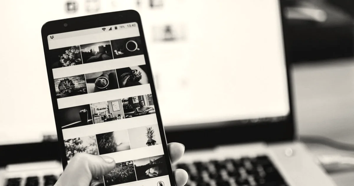 A person holding and browsing a smartphone displaying a social media feed in front of a laptop