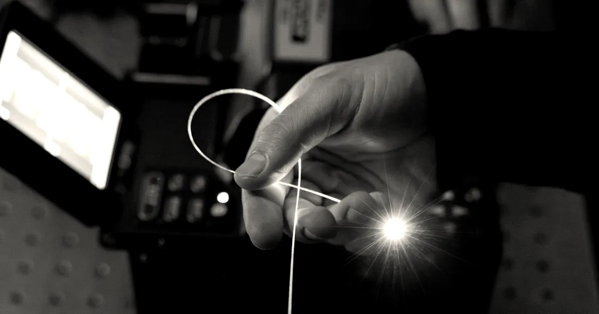 A hand holds a glowing strand of fiber optic cable emitting light at the tip in a dark lab.