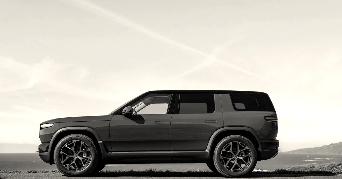 A side profile view of a dark green Rivian SUV parked near a coastal overlook under a clear sky.