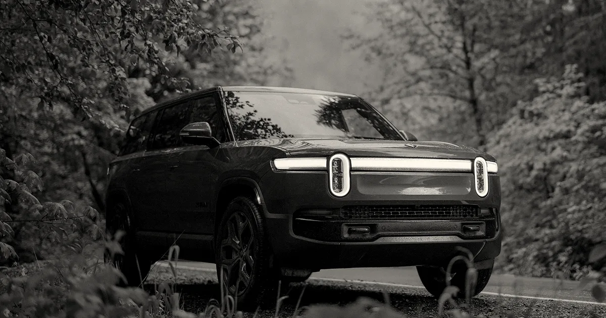 An orange Rivian R1S electric SUV driving on a road surrounded by lush green trees