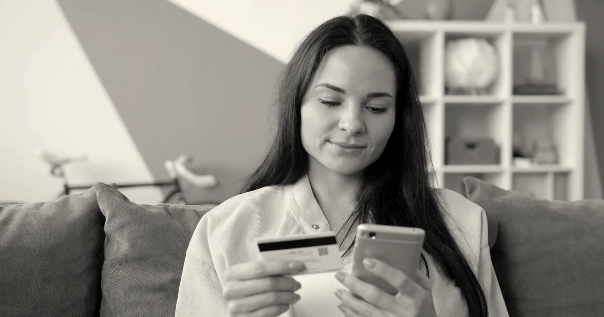 A woman holding a credit card while using a smartphone on a sofa.