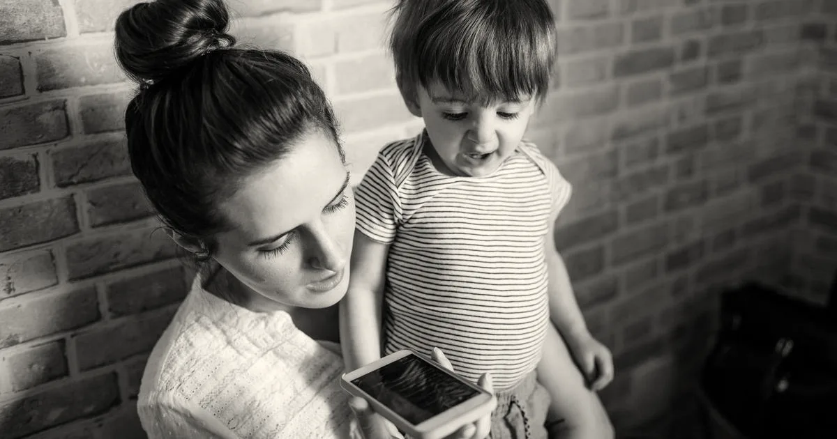 A mother holding a toddler while looking at a smartphone indoors.