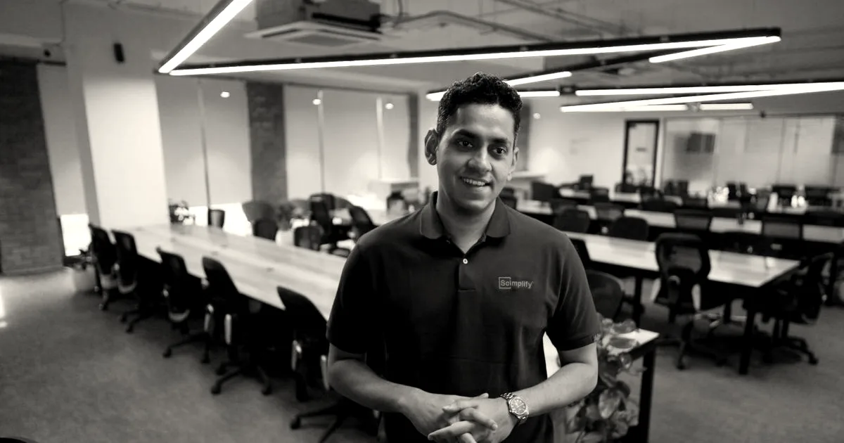 Scimplify co-founder Sachin Santhosh standing in an open-plan office space with hands clasped.