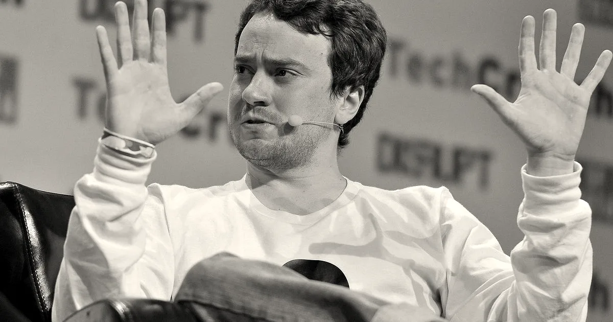 George Hotz speaking at TechCrunch Disrupt with his hands raised, wearing a white long-sleeve shirt and jeans.