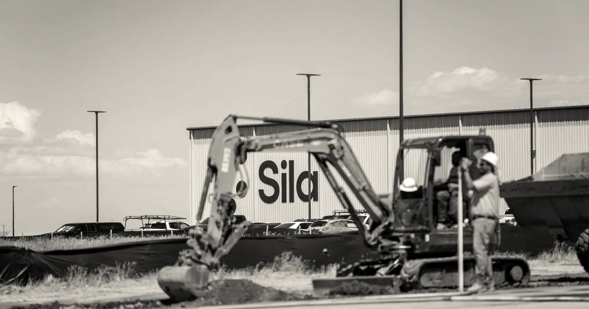 An excavator operating at the Sila facility with a worker standing nearby in front of a Sila building.