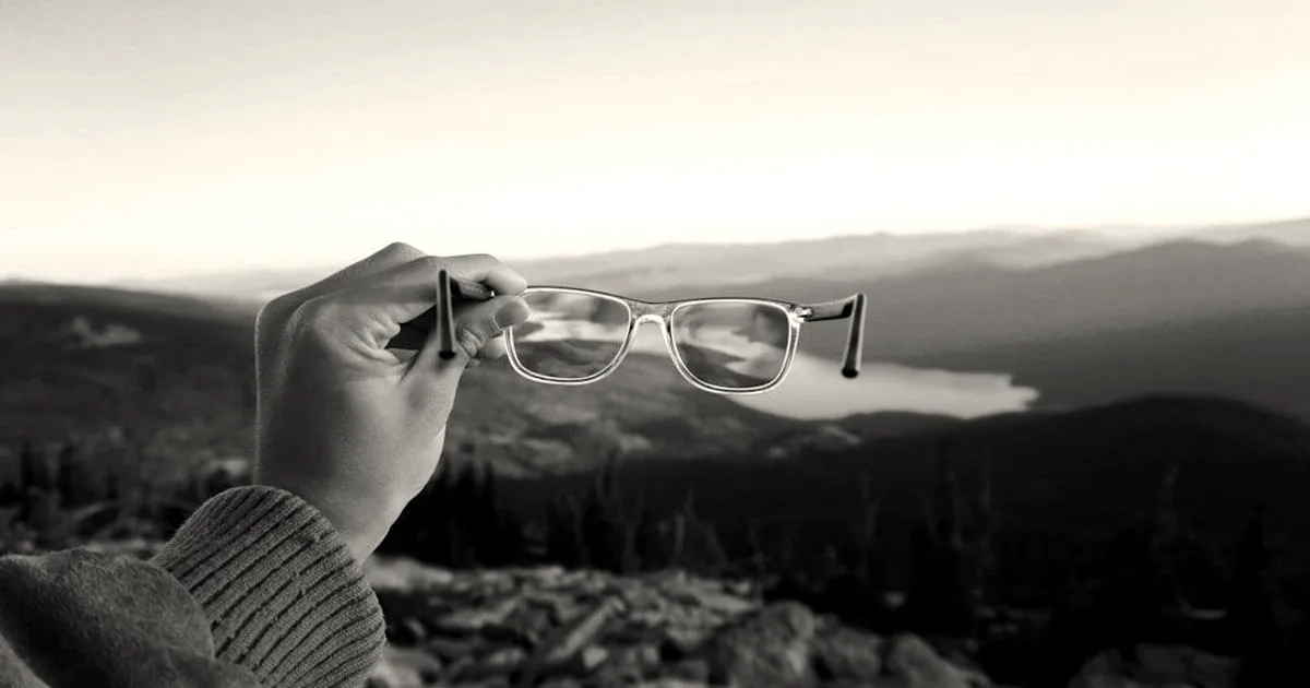 A person holding a pair of clear eyeglasses against a wide mountainous landscape