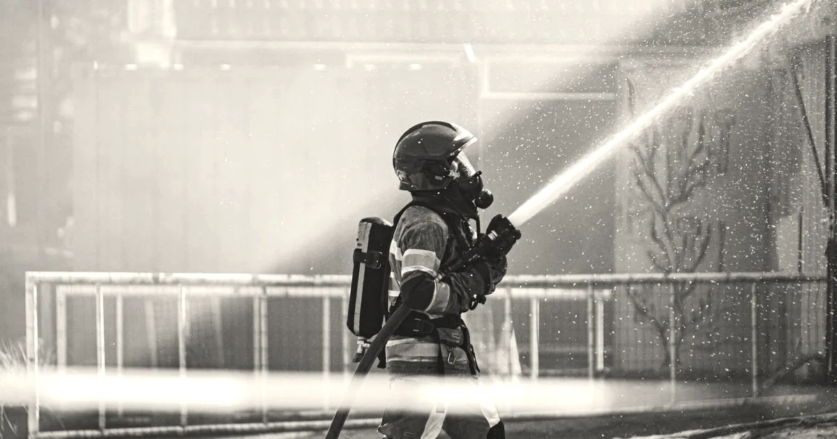 A firefighter in protective gear and red helmet spraying a water hose outdoors.