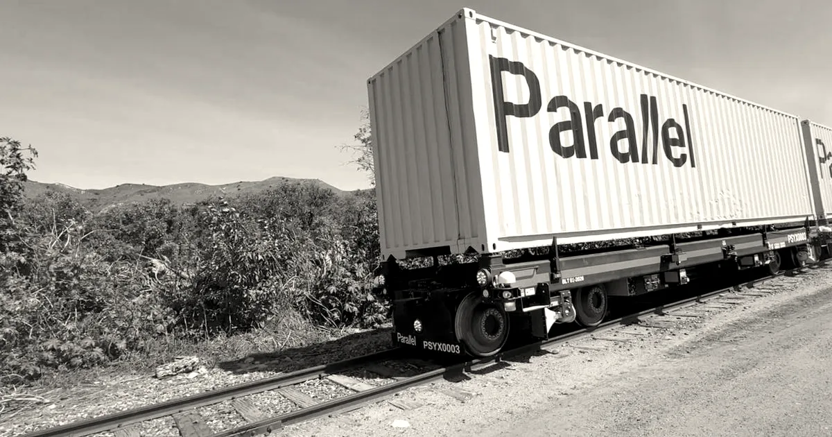 An autonomous electric freight rail vehicle carrying a Parallel shipping container along a track outdoors.