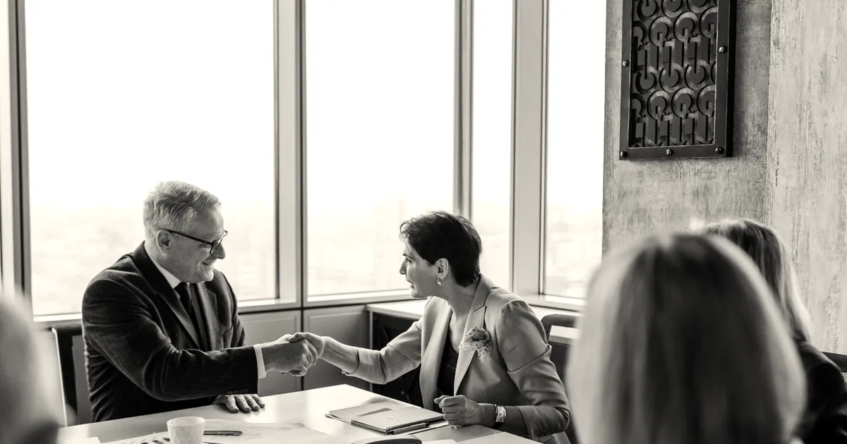 Two business executives in formal attire shaking hands across a conference table during a corporate meeting.