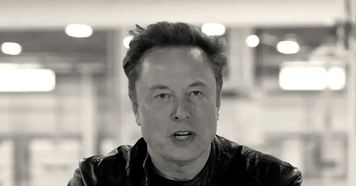 Elon Musk wearing a black leather jacket sitting at a white table speaking indoors.