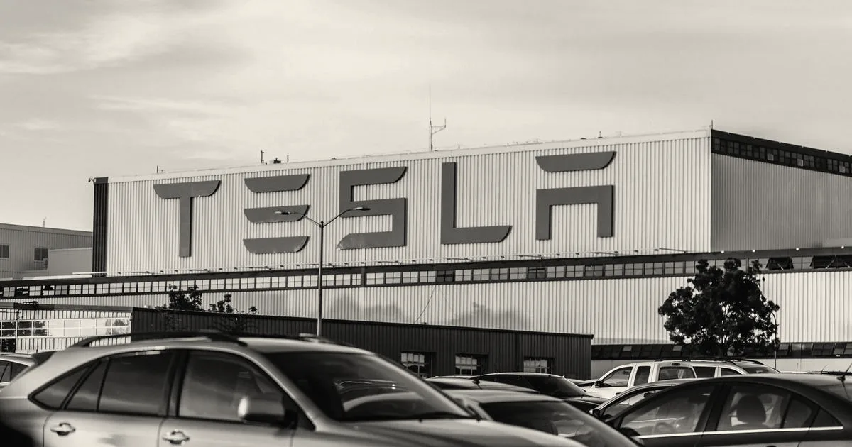 A Tesla factory building exterior with large red branding sign behind parked vehicles at sunset.