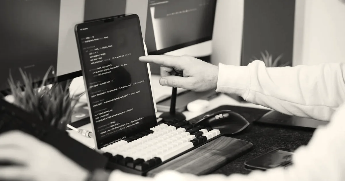A person pointing at lines of code displayed on a vertical monitor at a modern desk workstation
