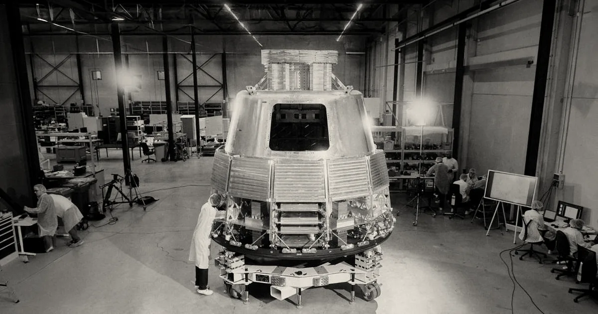 Engineers working on a spacecraft capsule prototype inside a spacious industrial assembly facility.