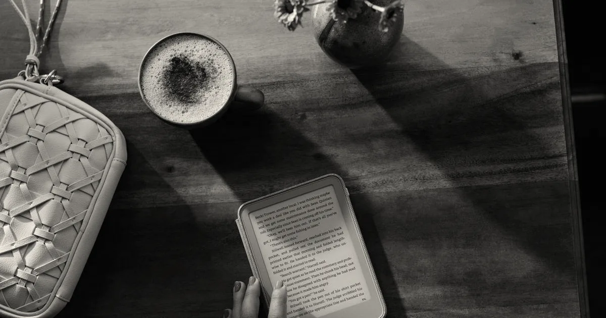 A person reading a purple Kindle on a wooden table beside a beverage and handbag