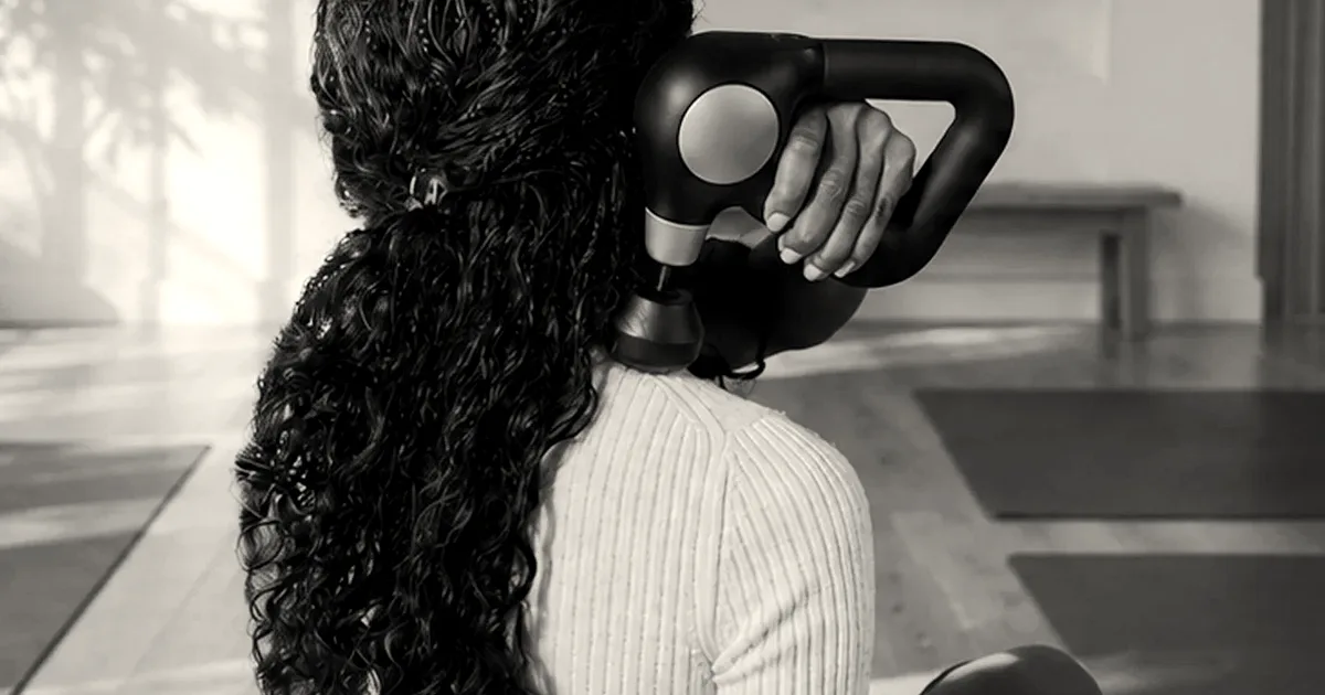 A woman using a black Theragun Sense massage device on her neck and shoulder area.