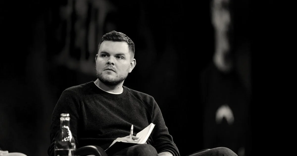 Man in dark sweater seated on stage holding a notebook and pen, listening intently
