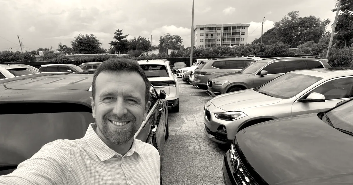 A smiling man takes a selfie outdoors in a parking lot filled with parked cars.