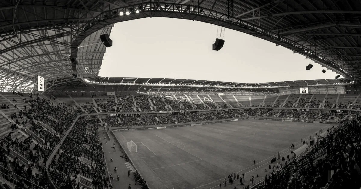 Wide panoramic view of a soccer stadium packed with fans during a match under a curved canopy.