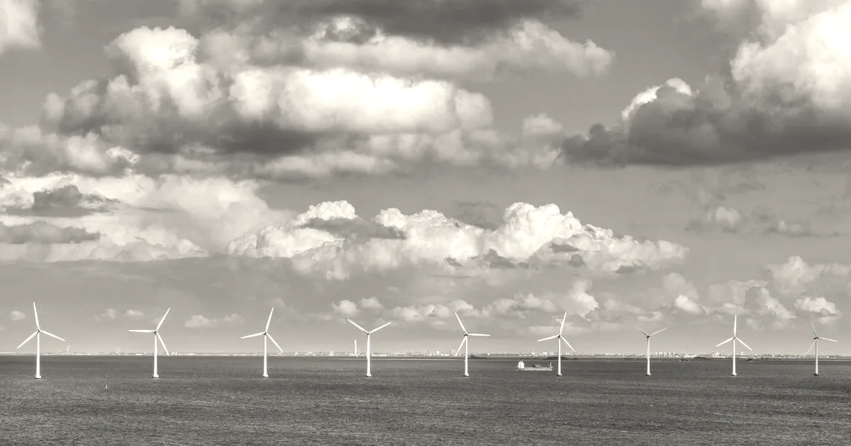 A line of offshore wind turbines standing in the open sea under a cloudy blue sky.