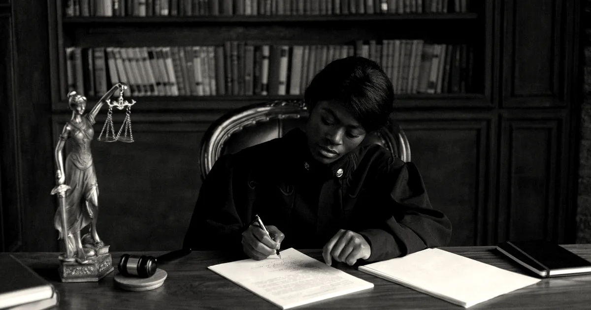 A judge in robes writing on a document at a desk in an office library with law books.