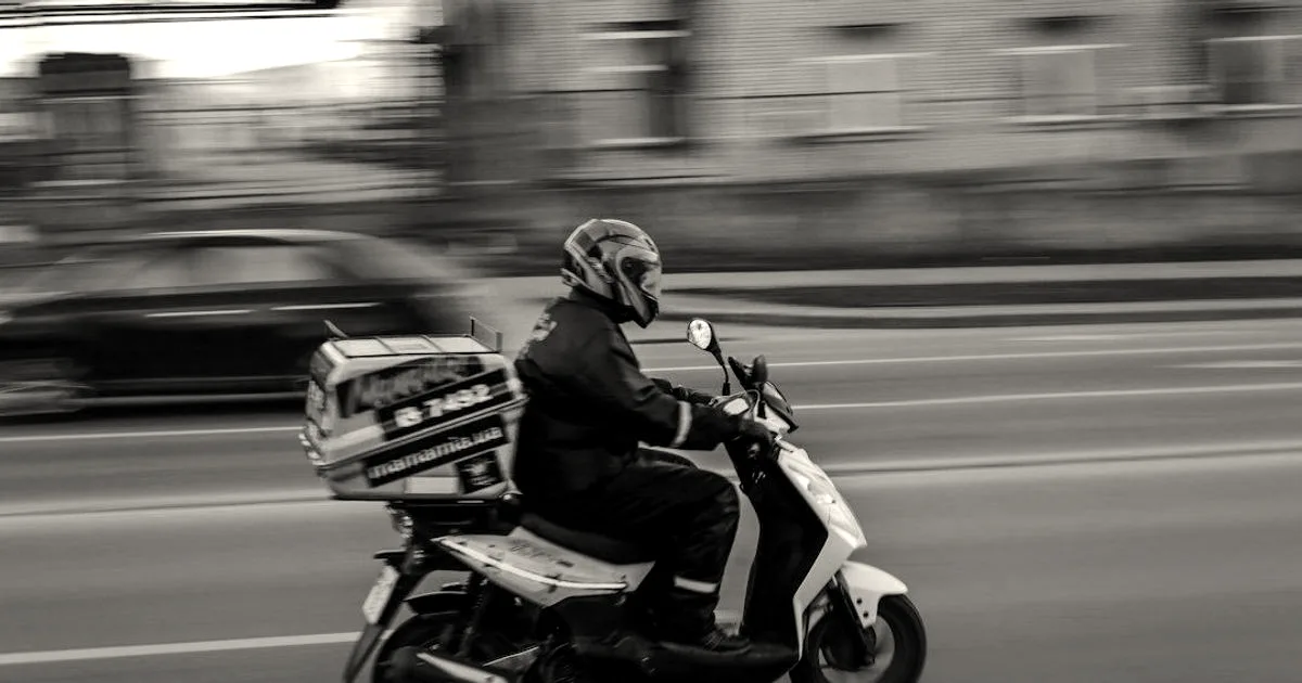 A delivery rider wearing a helmet and jacket on a moving scooter with a rear cargo box