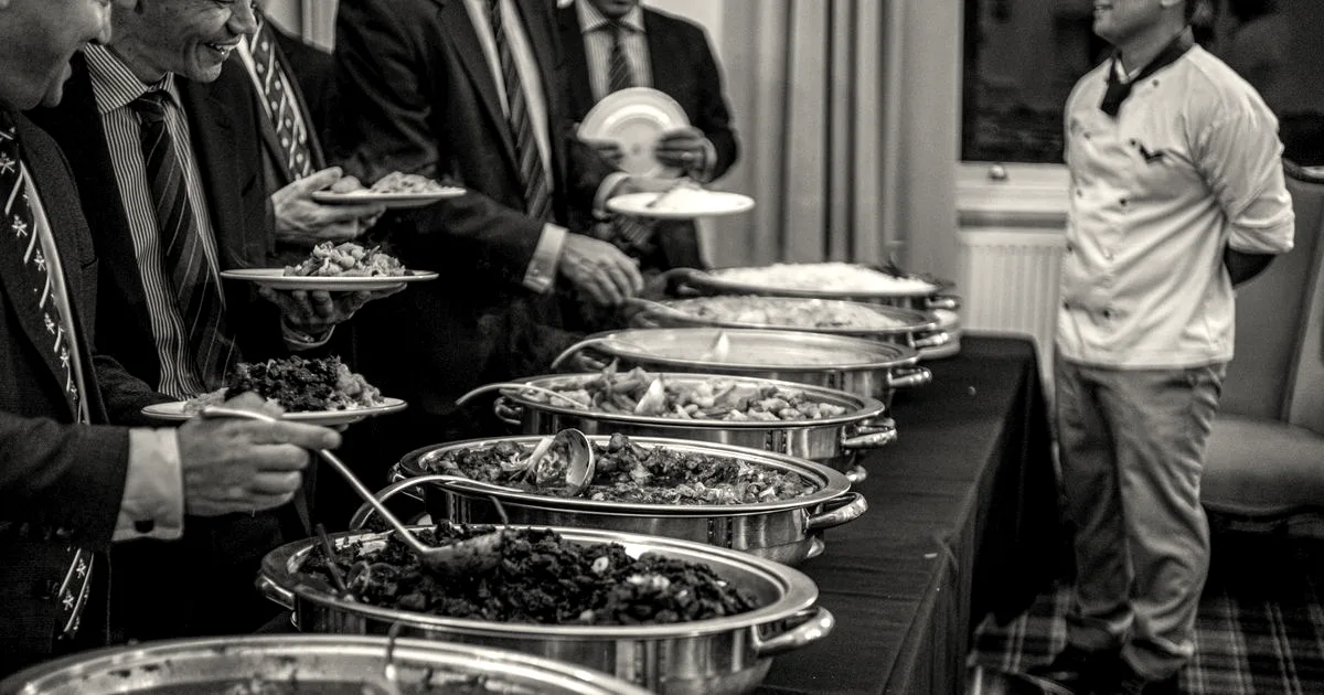 Professionals in business suits serving food from a catered buffet line with chafing dishes.