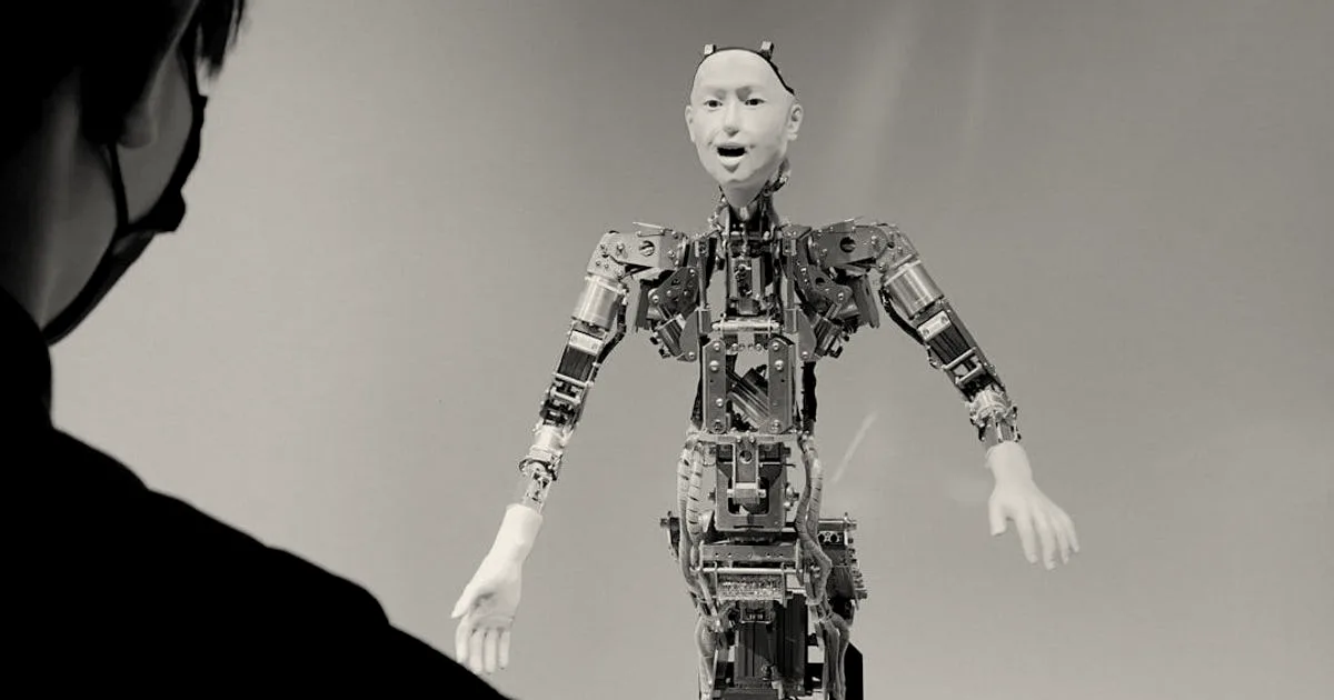 A person standing on the left looks at a metallic humanoid robot on a grey background.