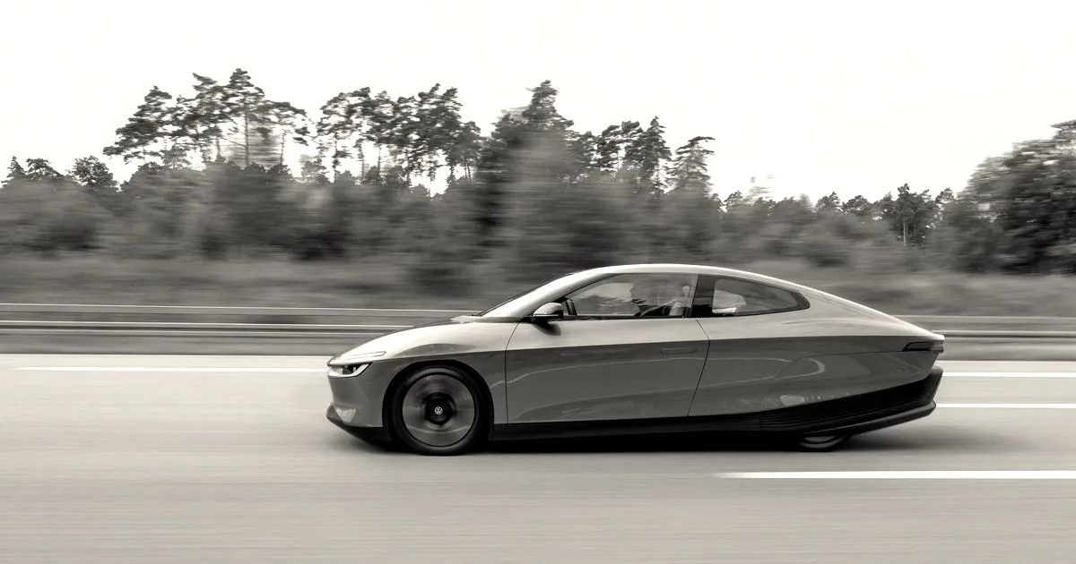 A sleek, silver aerodynamic Volkswagen prototype electric vehicle driving along a paved road past trees.